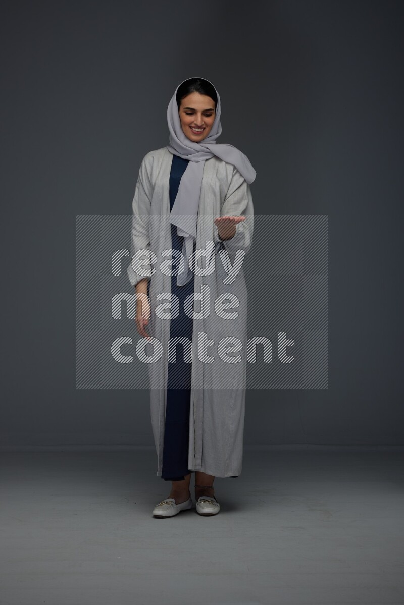A Saudi woman wearing a light gray Abaya and head scarf standing and pointing in different directions eye level on a grey background