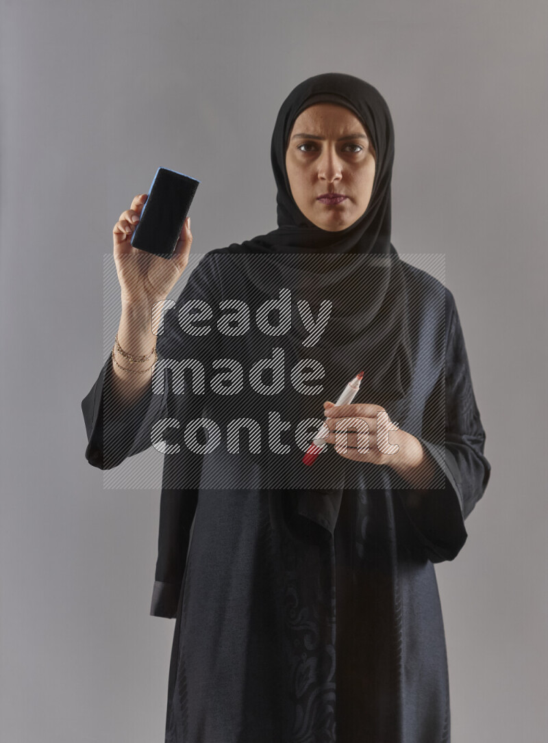 Woman in black abaya and hijab holding a marker pen to write on a transparent board with different reactions and expressions on grey background