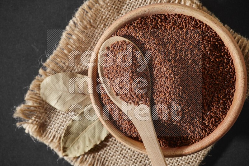 A wooden bowl full of garden cress seeds with wooden spoon full of the seeds on it on burlap fabric on a black flooring