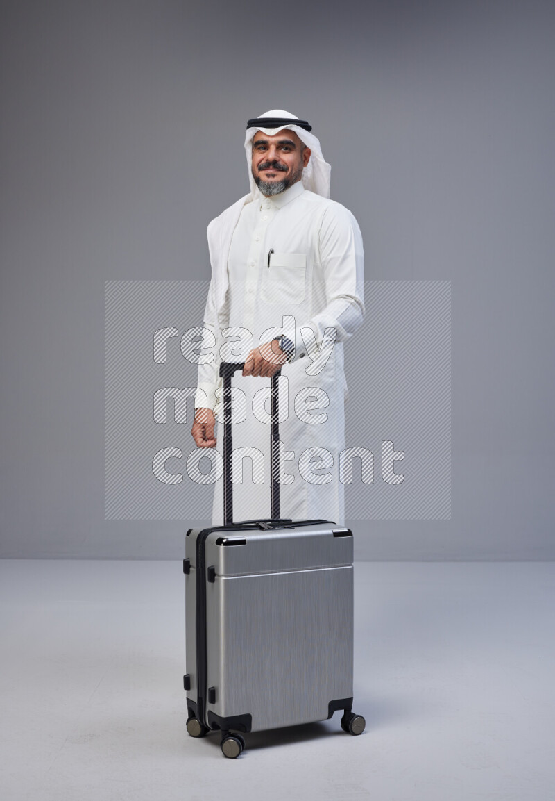 Saudi man wearing Thob and white Shomag standing holding Travel bag on Gray background