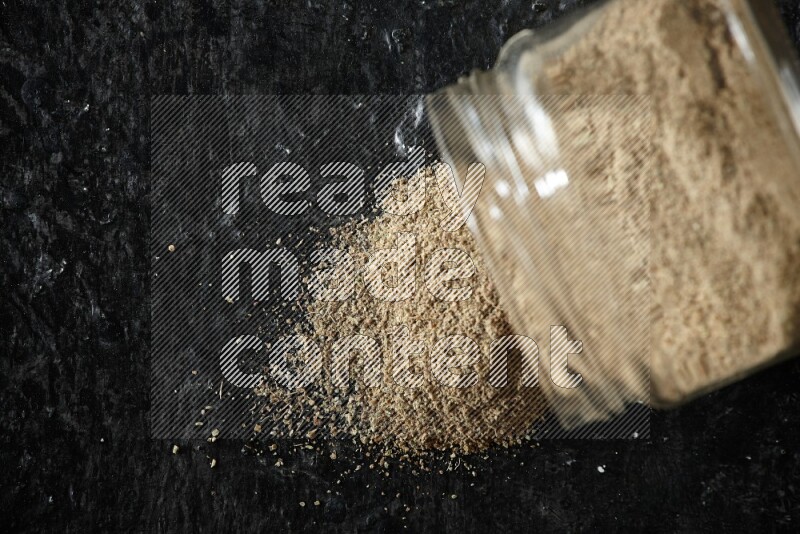 A flipped glass jar full of cardamom powder and powder spilled out of it on textured black flooring