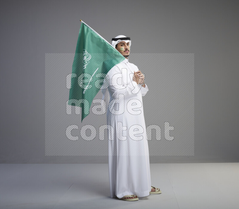 A Saudi man standing wearing thob and white shomag with face painting raising big Saudi flag on gray background