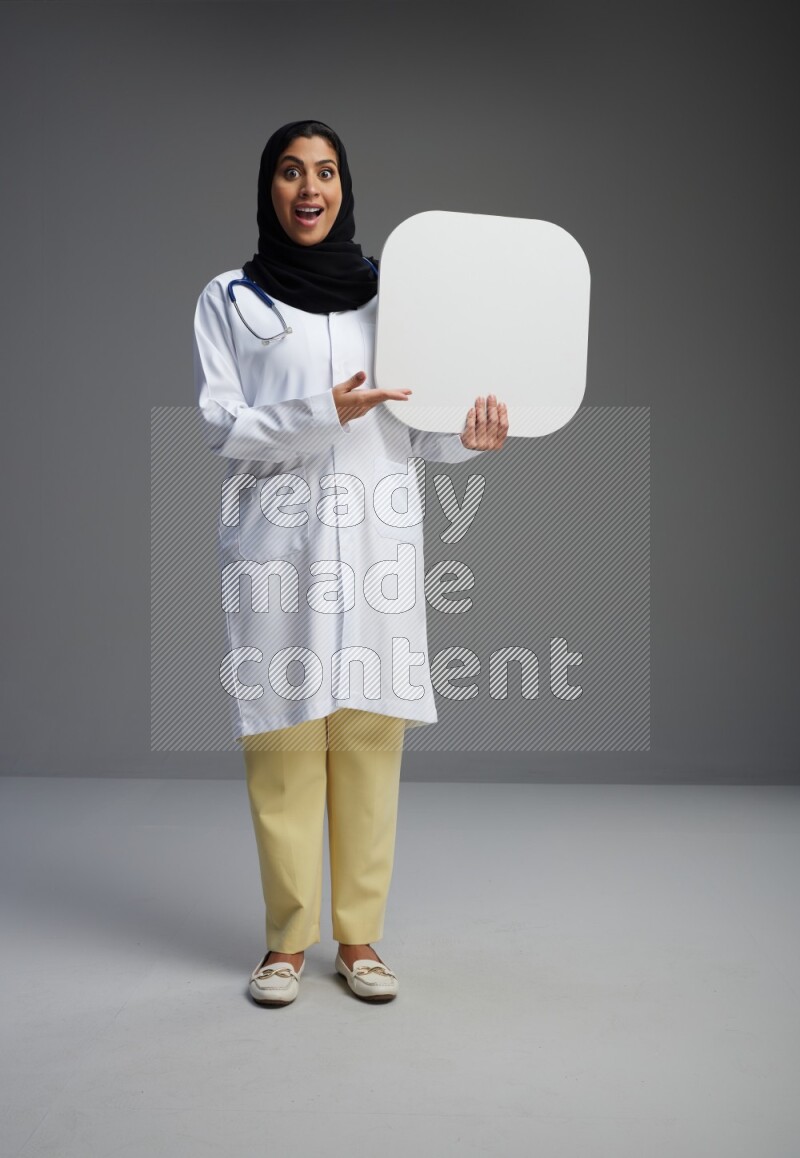 Saudi woman wearing lab coat with stethoscope standing holding social media sign on Gray background