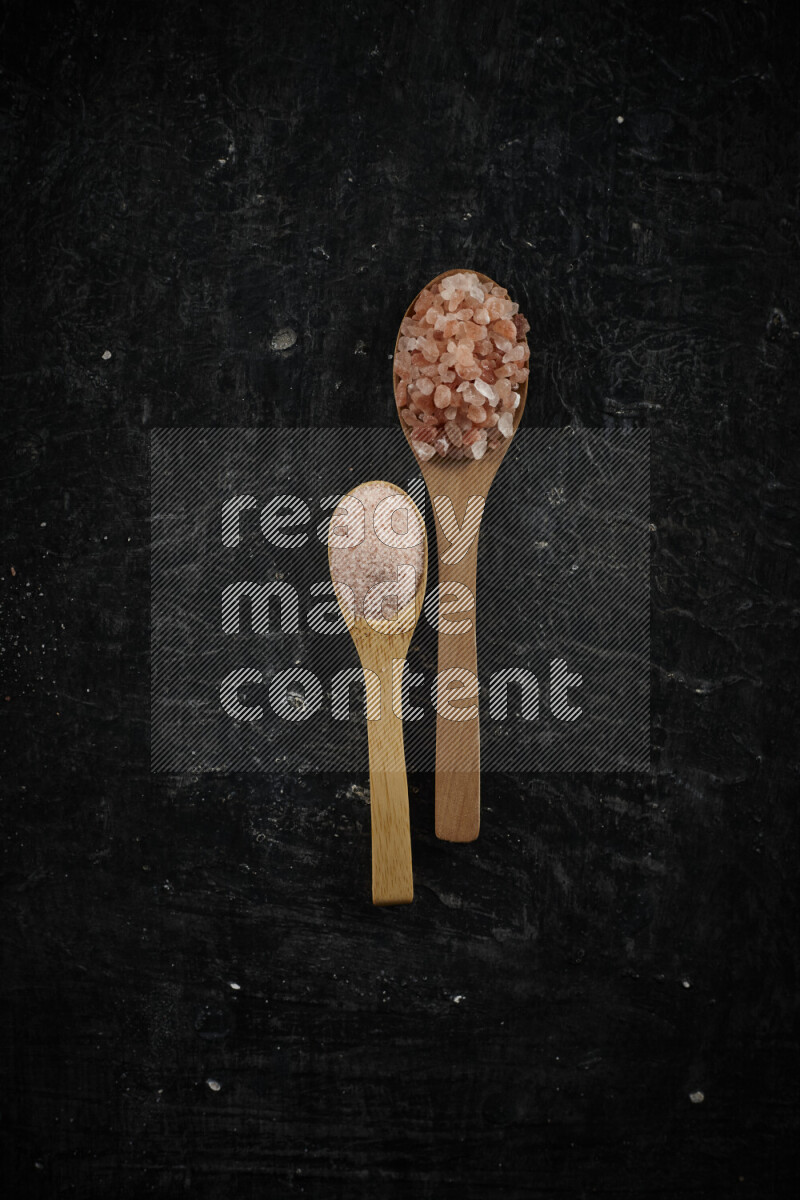 2 wooden spoons filled with fine and coarse salt on black background