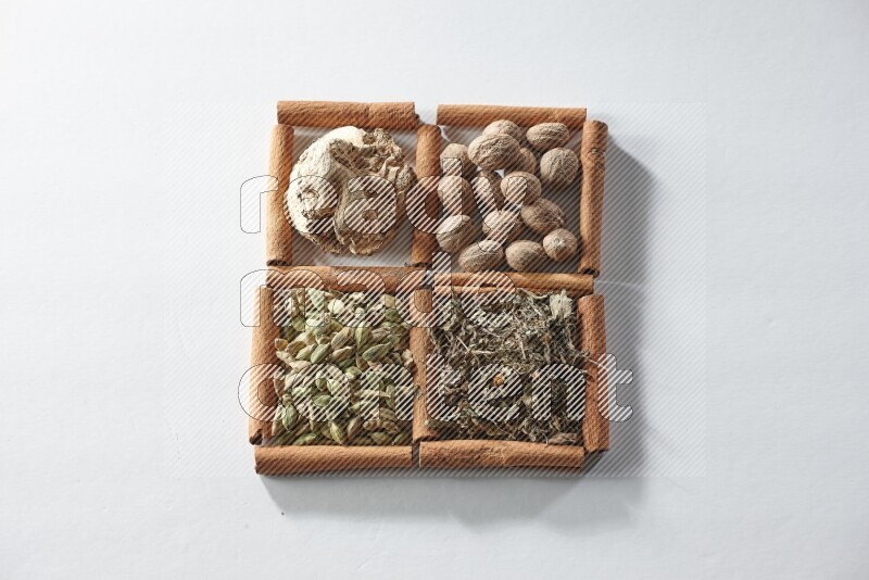 4 مربعات من أعواد القرفة ممتلئة بجوزة الطيب والزنجبيل المجفف والهيل والريحان المجفف على أرضية بيضاء