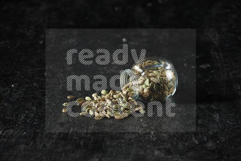 A flipped glass spice jar full of cardamom seeds on textured black flooring