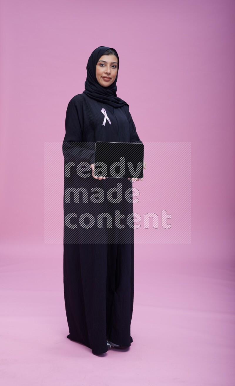 Saudi woman wearing pink ribbon on Abaya standing showing a tablet to camera on pink background