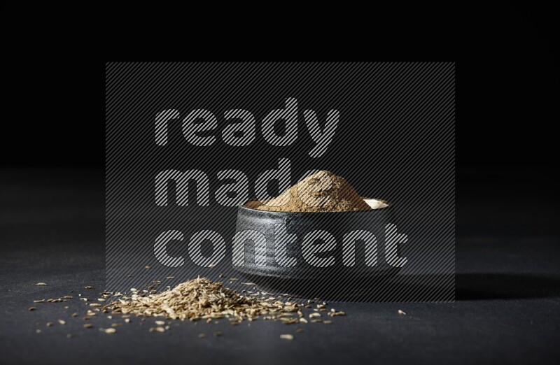 وعاء فخاري أسود ممتلئ ببودرة الكمون وحبوب الكمون متناثرة على خلفية سوداء