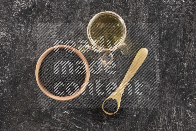 A wooden bowl and spoon full of black seeds and a glass jar of black seeds oil on a textured black flooring