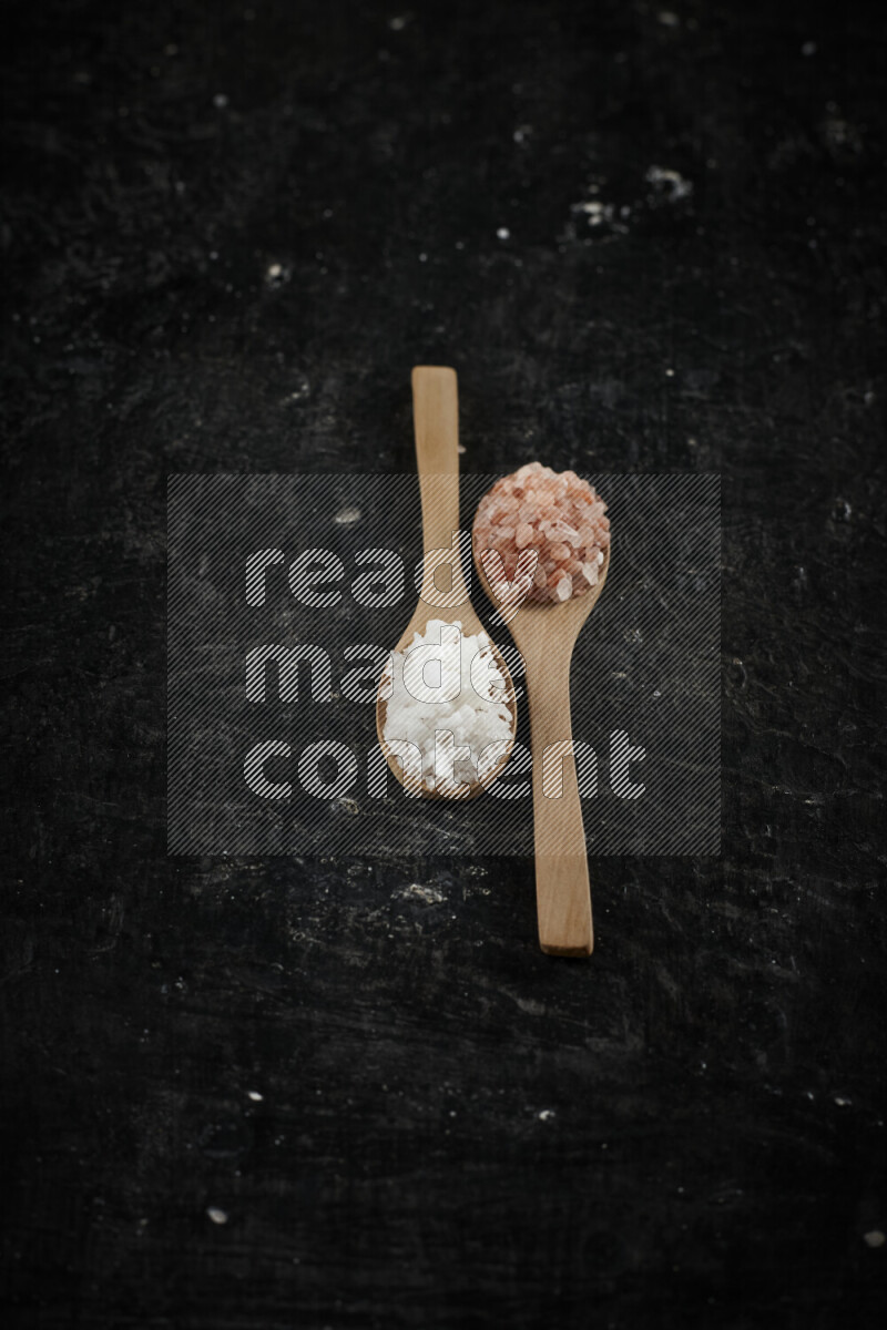 2 wooden spoons filled with fine and coarse salt on black background