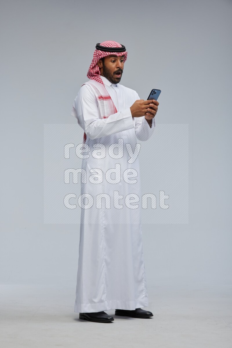 Saudi man Wearing Thob and shomag standing texting on phone on Gray background