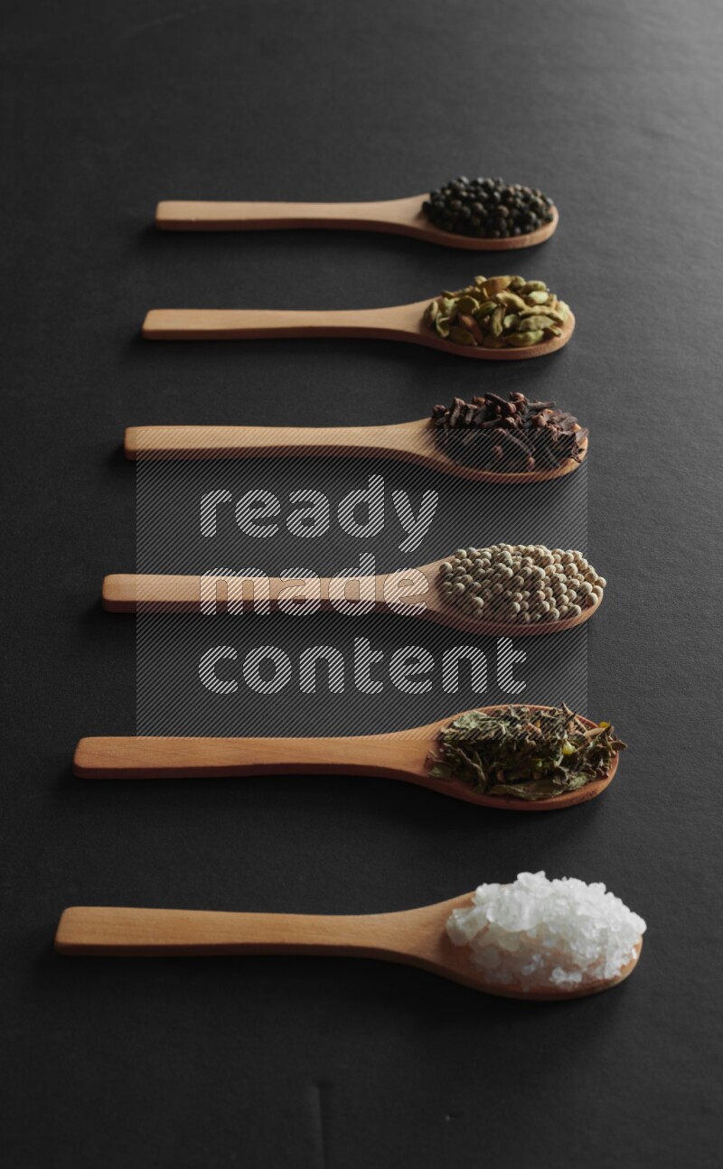 wooden spoons filled with white peppers, cloves, cardamom, salt, black peppers and basil on black flooring
