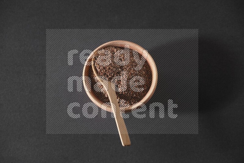 A wooden bowl full of flaxseeds with wooden spoon full of the seeds on it on a black flooring