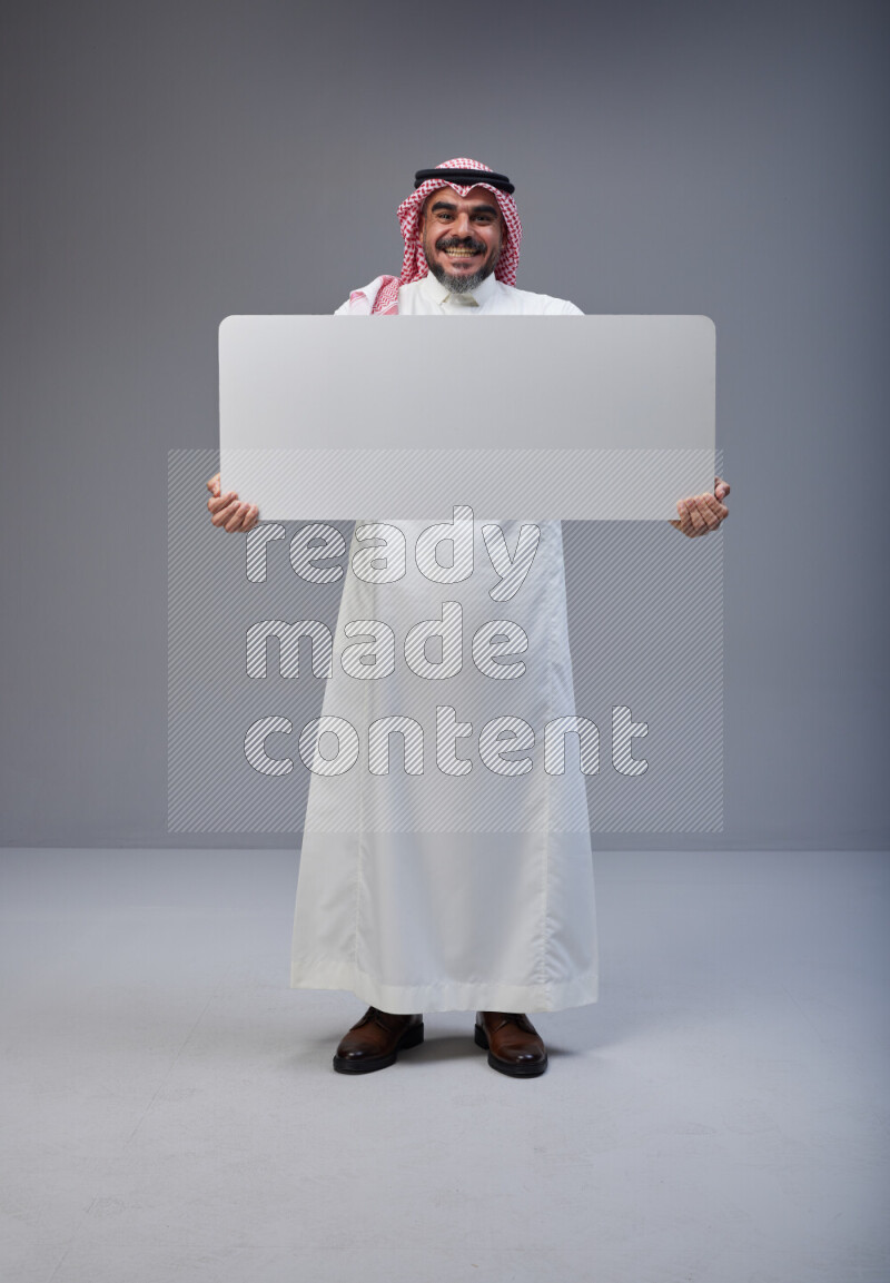 Saudi man Wearing Thob and red Shomag standing holding board on Gray background