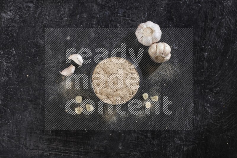 A black pottery bowl full of garlic powder with garlic cloves and a whole bulb beside it on a textured black flooring