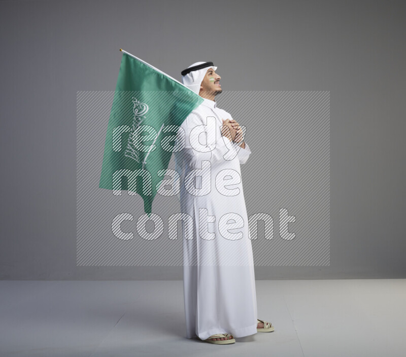 A Saudi man standing wearing thob and white shomag with face painting raising big Saudi flag on gray background