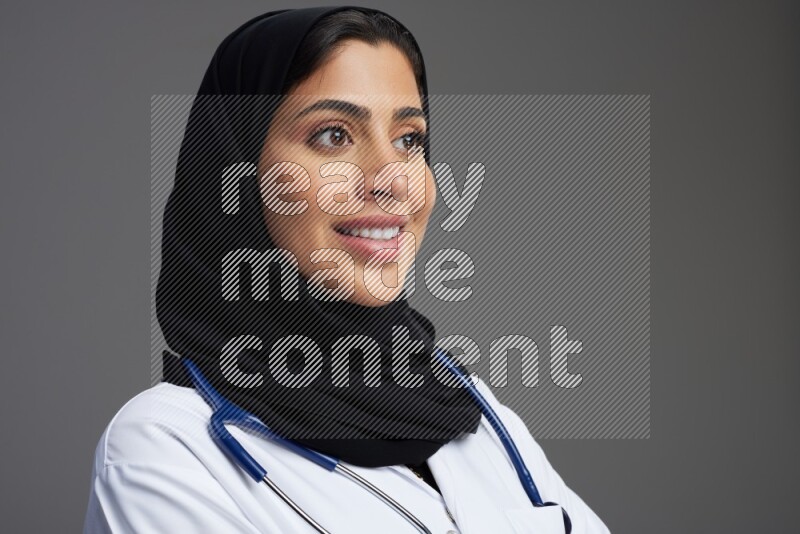Saudi woman wearing lab coat with stethoscope standing with crossed arms on Gray background