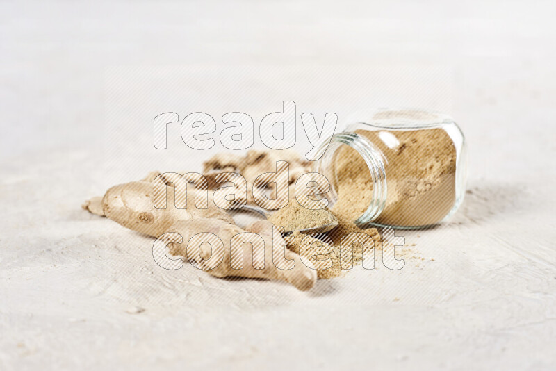 A glass jar full of ground ginger powder flipped with some spilling powder on white background