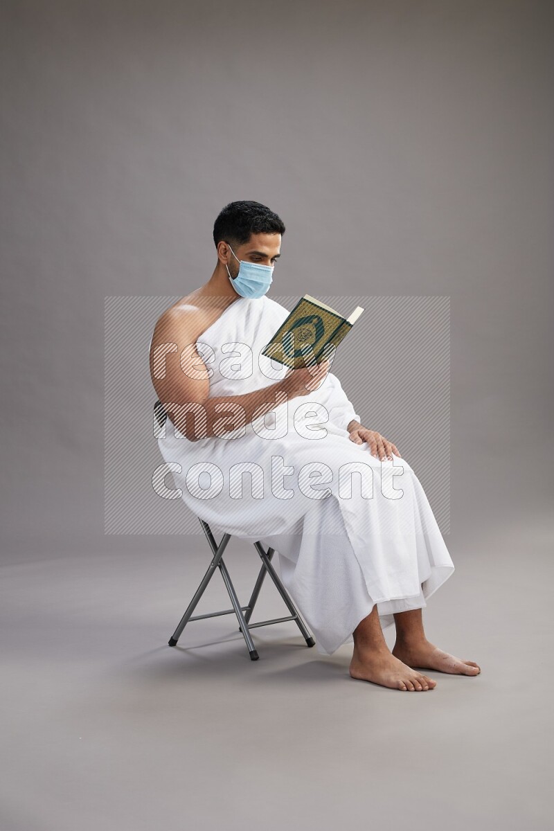 A man wearing Ehram with face mask sitting on chair reading quran on gray background