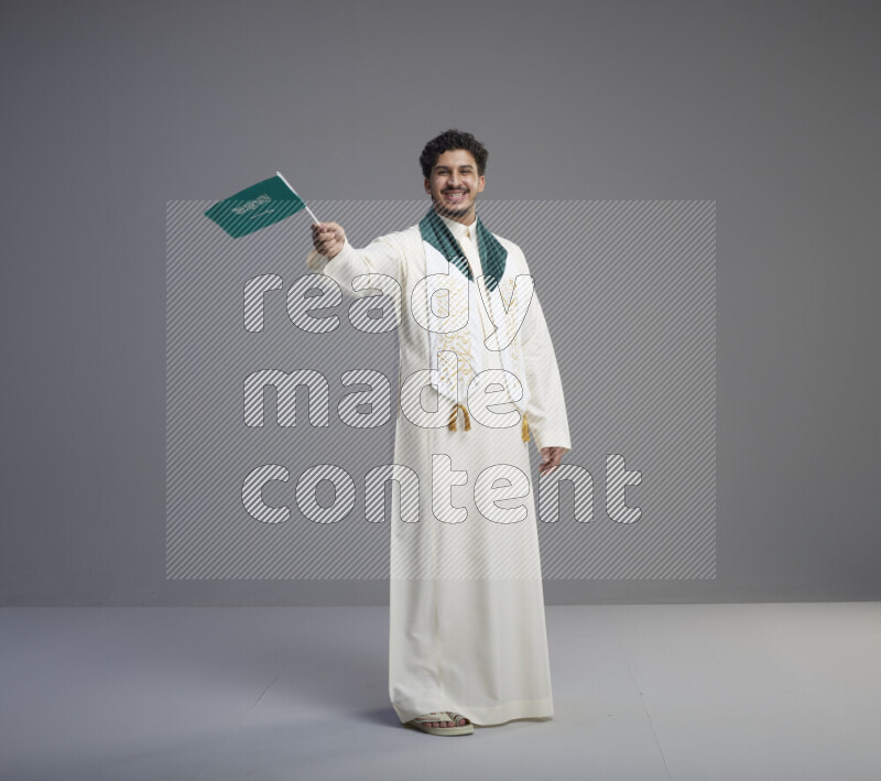 A Saudi man standing wearing thob and saudi flag scarf and holding small saudi flag on gray background