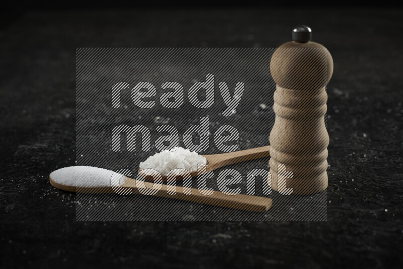 2 wooden spoons filled with fine and coarse salt with wooden grinder beside them on black background