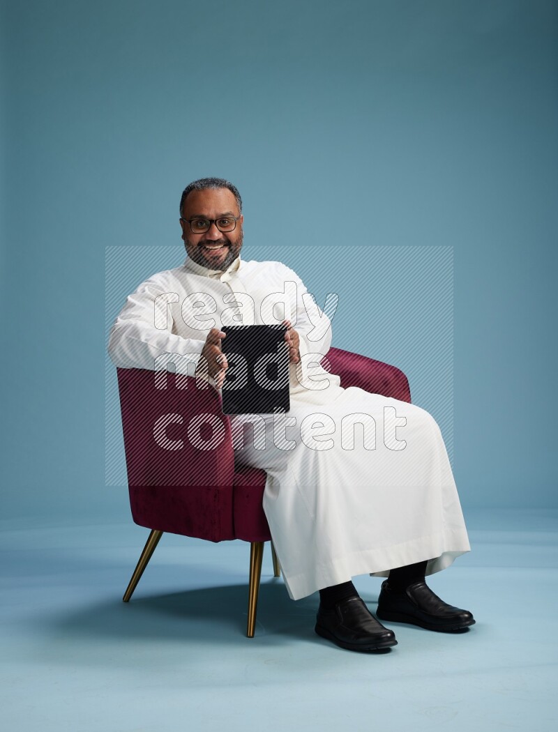Saudi Man without shimag sitting on chair working on tablet on blue background