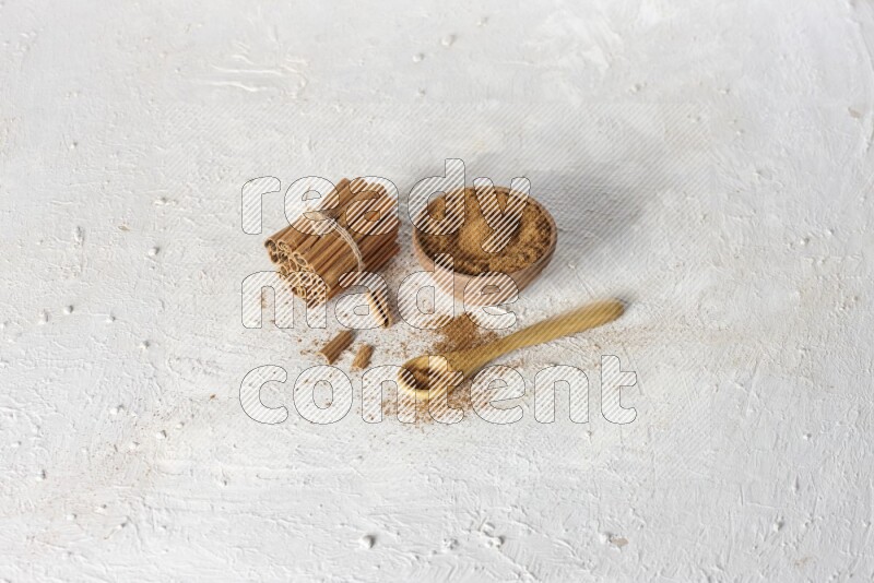 Cinnamon sticks stacked and bounded beside a wooden bowl full of cinnamon powder and a wooden spoon full of powder on white background