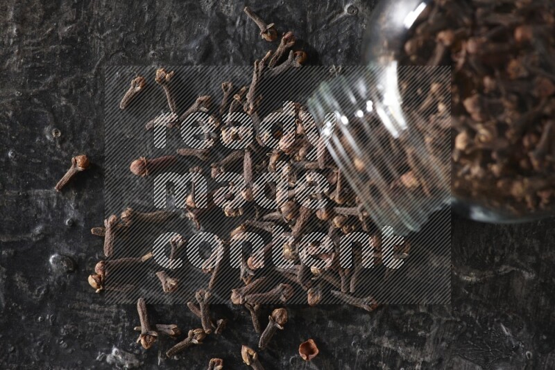 وعاء زجاجي للبهارات مقلوب ممتلئ بالقرنفل والقرنفل سقط منه على أرضية سوداء