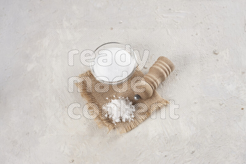 A glass bowl full of white salt with a wooden grinder on a burlap fabric all on white background