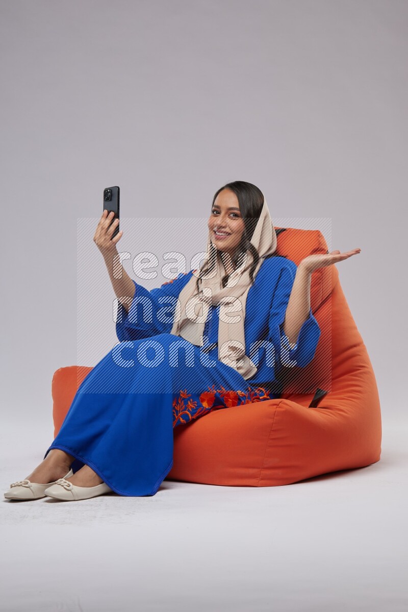 A Saudi woman wearing jalabiya sitting on an orange beanbag and taking selfie