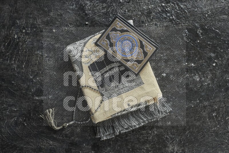 A folded prayer mat with different elements such as quran and prayer beads on black textured background