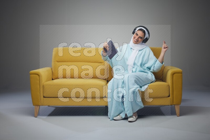 A Saudi woman wearing a light blue Abaya and a white head scarf sitting on a yellow sofa and holding an iPad while wearing headphone eye level on a grey background