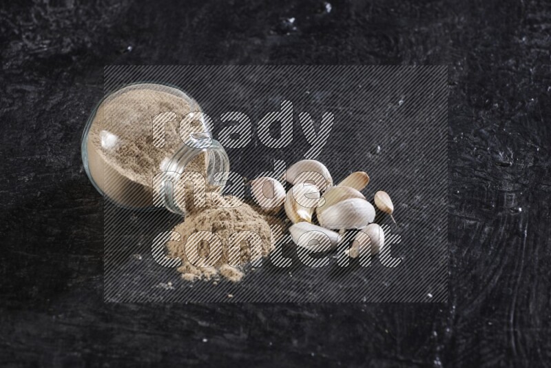 A glass spice jar full of garlic powder flipped over with the powder came out on a textured black flooring
