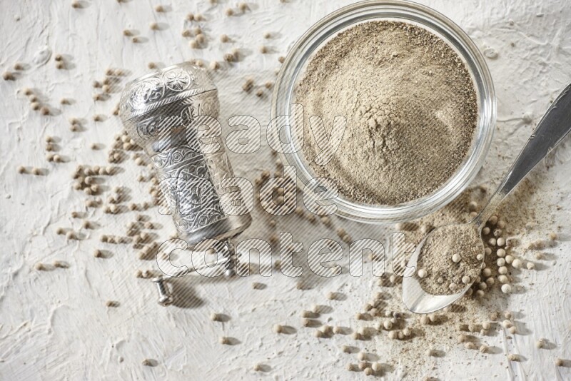 A glass bowl full of white pepper powder with white pepper beads and a metal grinder on textured white flooring