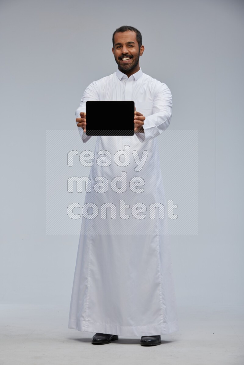 Saudi man wearing Thob standing showing tablet to camera on Gray background