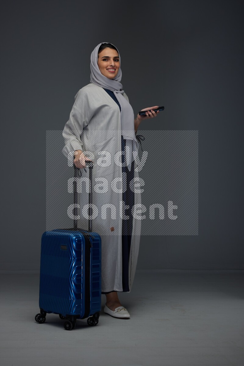 A Saudi woman wearing a light gray Abaya and head scarf standing holding a phone and being shocked  eye level on a grey background