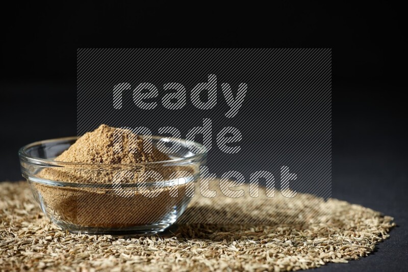 A glass bowl full of cumin powder surrounded by cumin seeds on black flooring