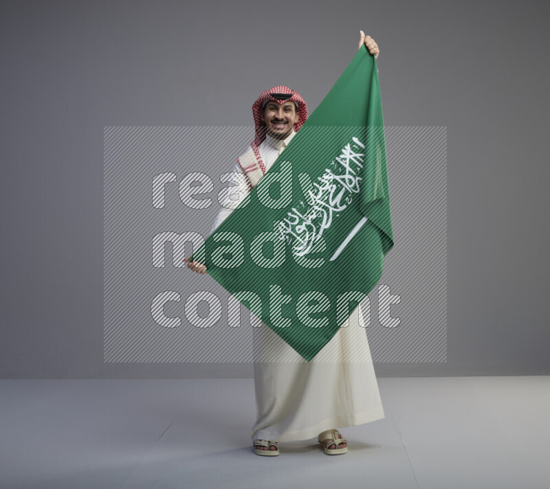A saudi man standing wearing thob and red shomag holding big saudi flag on gray background