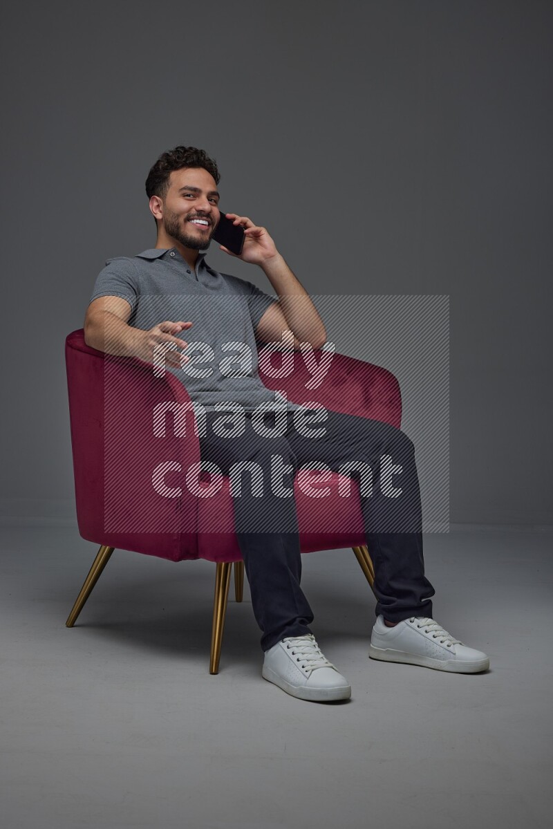 A man wearing casual and talking in his phone while setting on a burgundy chair eye level on a gray background