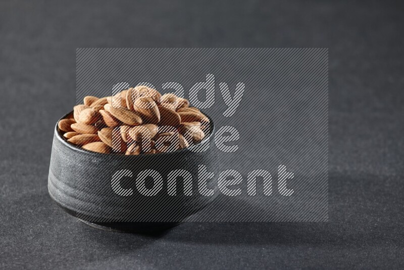 A black pottery bowl full of peeled almonds on a black background in different angles
