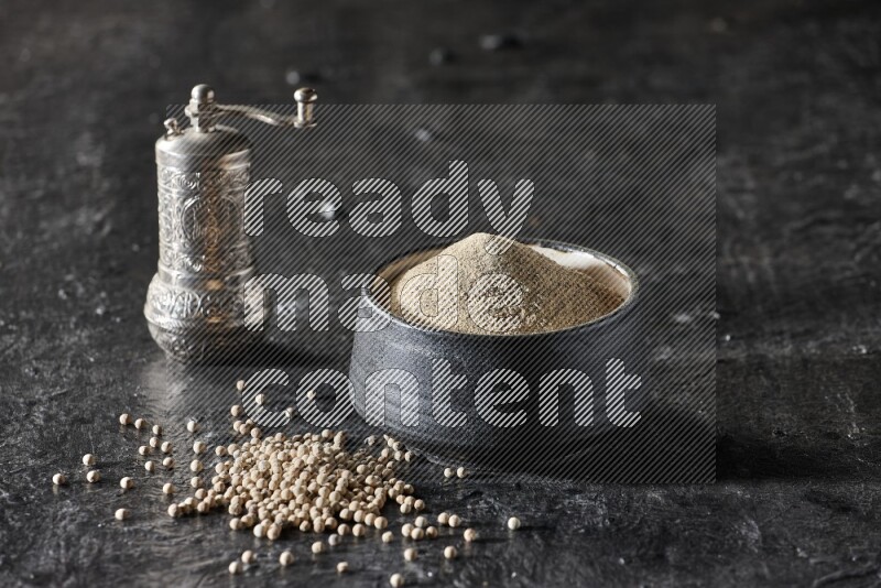 Black pottery bowl full of white pepper powder with pepper beads and metal pepper grinder on textured black flooring