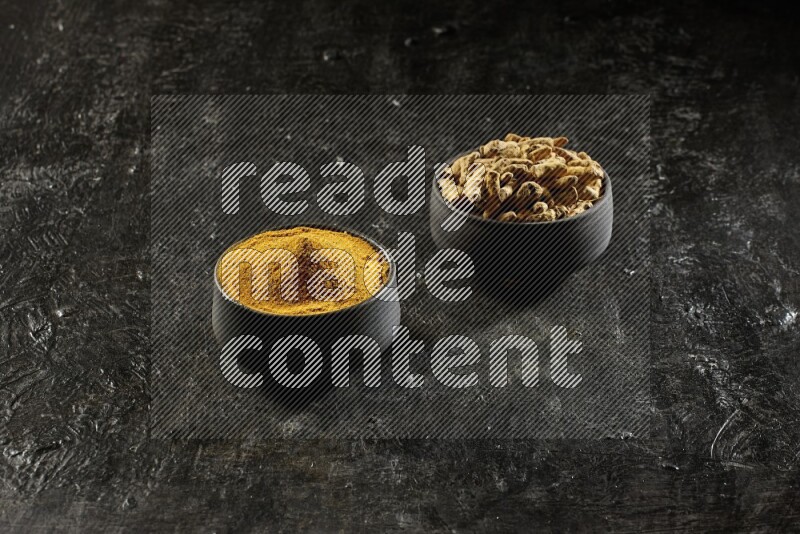 2 pottery black bowls, one full of turmeric powder and the other full of dried turmeric whole fingers on a textured black flooring