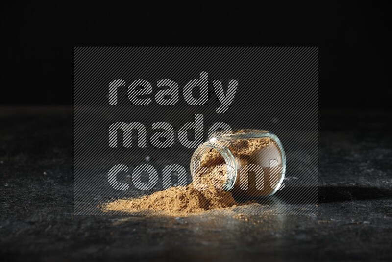 A flipped glass spice jar full of allspice powder and powder spilled out of it on a textured black flooring