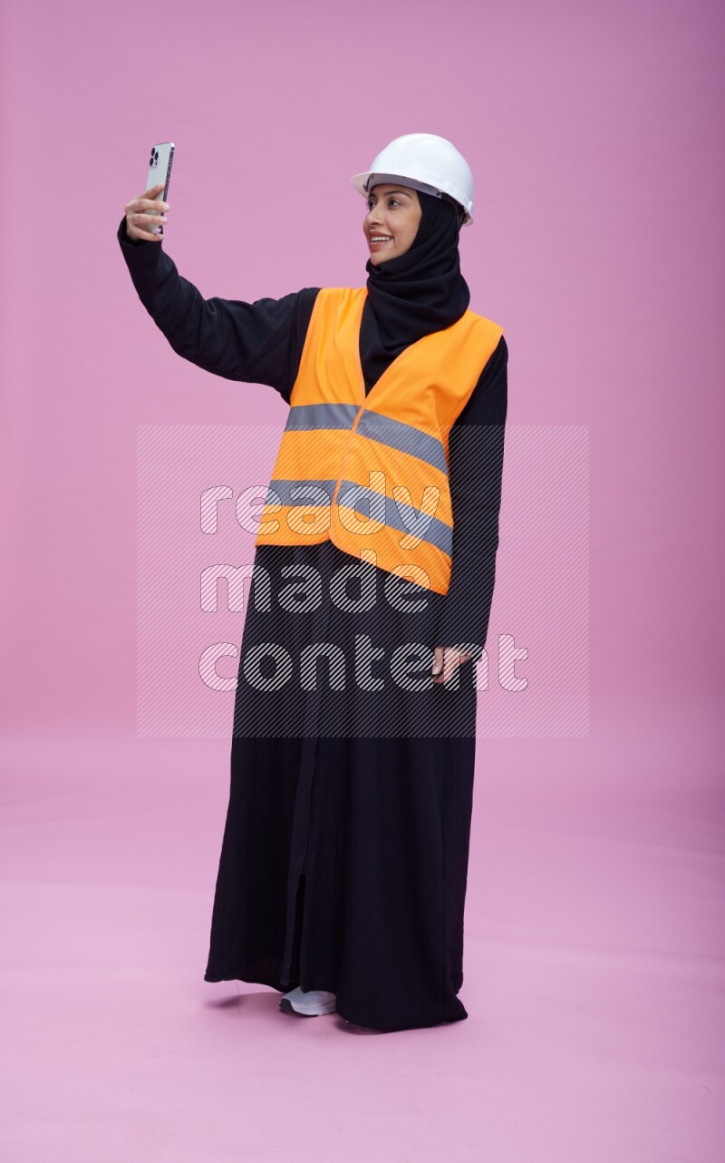 Saudi woman wearing Abaya with engineer vest and helmet standing taking selfie on pink background