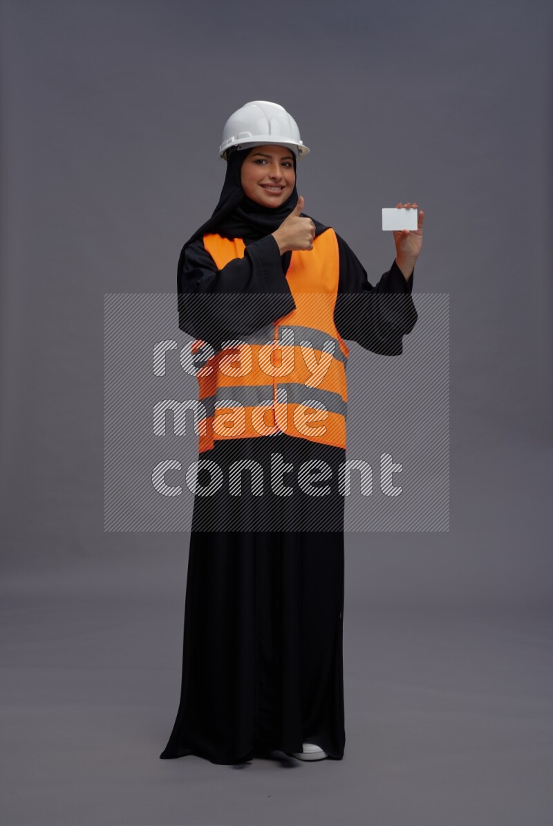 Saudi woman wearing Abaya with engineer vest standing holding ATM card on gray background