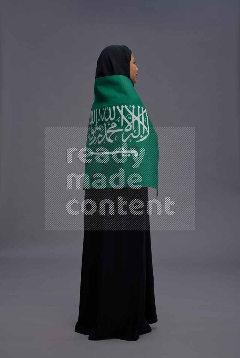 Saudi woman wearing Abaya standing holding Saudi flag on gray background