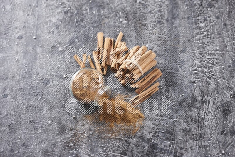 Herbal glass jar full of cinnamon powder flipped with cinnamon sticks stacked and bounded on a textured black background