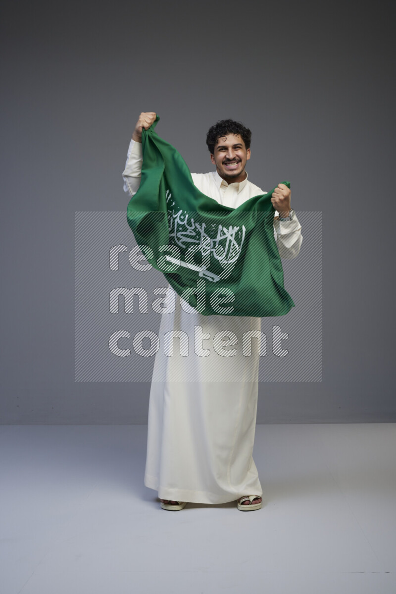 A Saudi man standing wearing thob holding big Saudi flag on gray background
