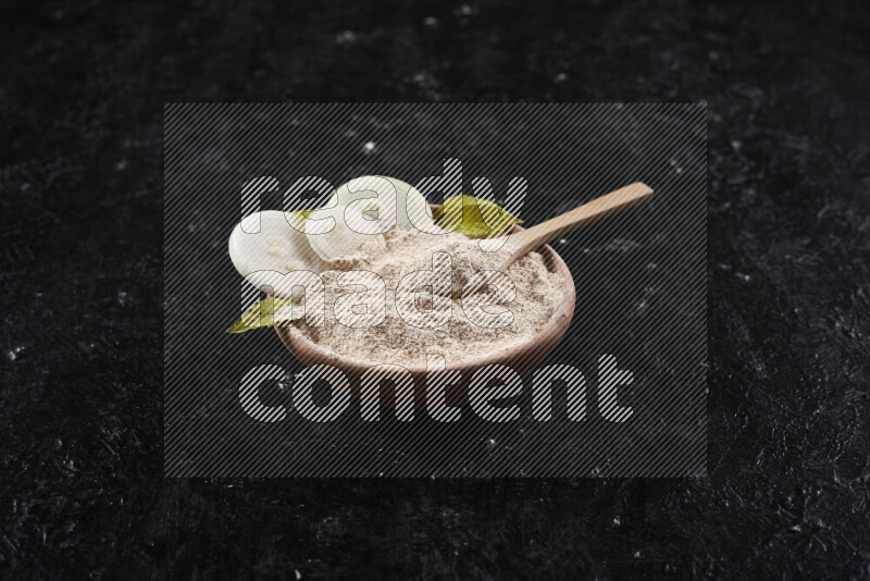 A wooden bowl full of onion powder with a wooden spoon in it with some sliced onions on black background
