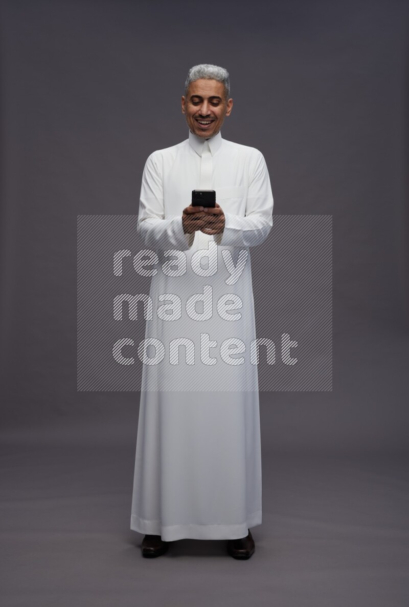 Saudi man wearing thob standing texting on phone on gray background